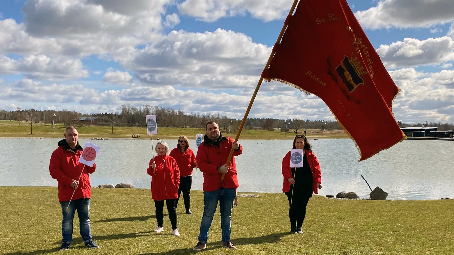 Tre personer i röda jackor och en stor röd flagga vid en sjö.