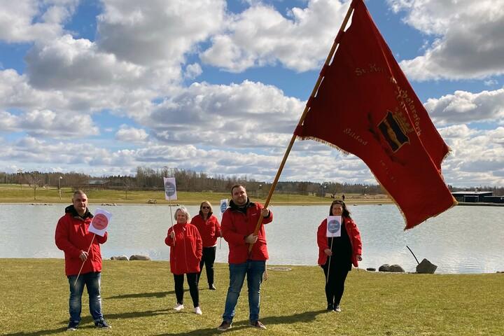 Tre personer i röda jackor och en stor röd flagga vid en sjö.